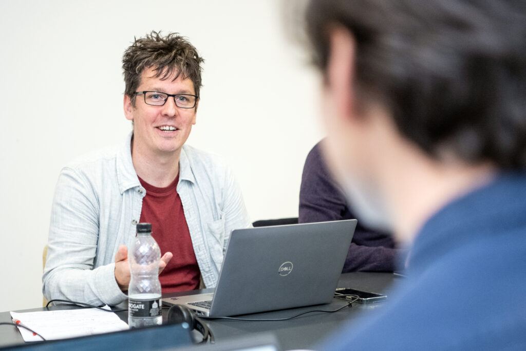 Assistive technology. A Blazie trainer is seated at a table, smiling while speaking. Another person, slightly out of focus, is in the foreground, facing him. The setting is a training session.